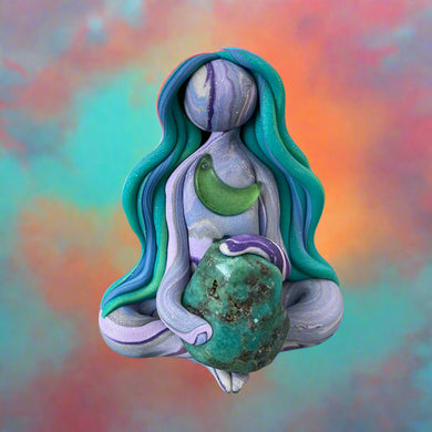 Colorful polymer clay figurine of a woman with long hair and a green stone on a decorative surface.